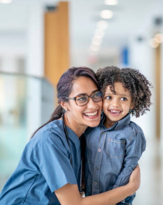 A nurse embracing a happy young child in a hospital hallway.