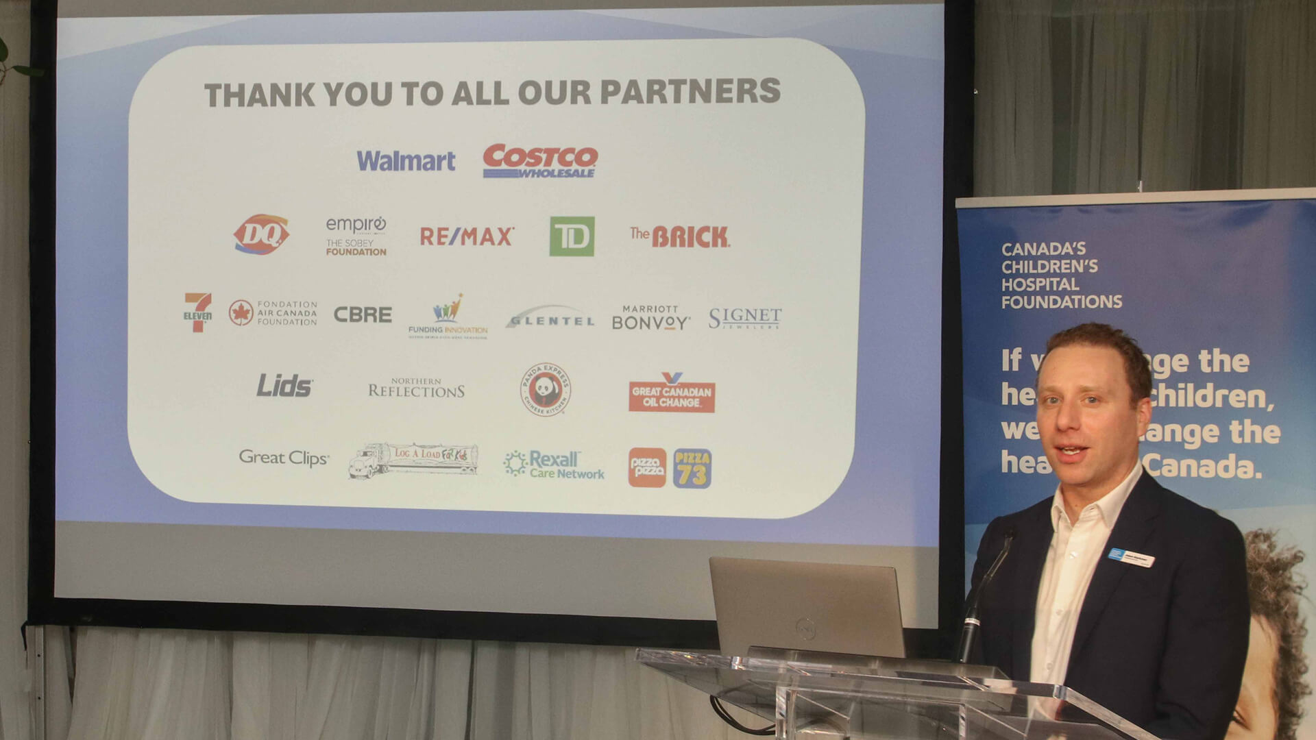 Le PDG du CCHF, Adam Starkman, debout à côté de l'écran, remercie les commanditaires d'entreprise comme Walmart, TD, RE/MAX et Dairy Queen.